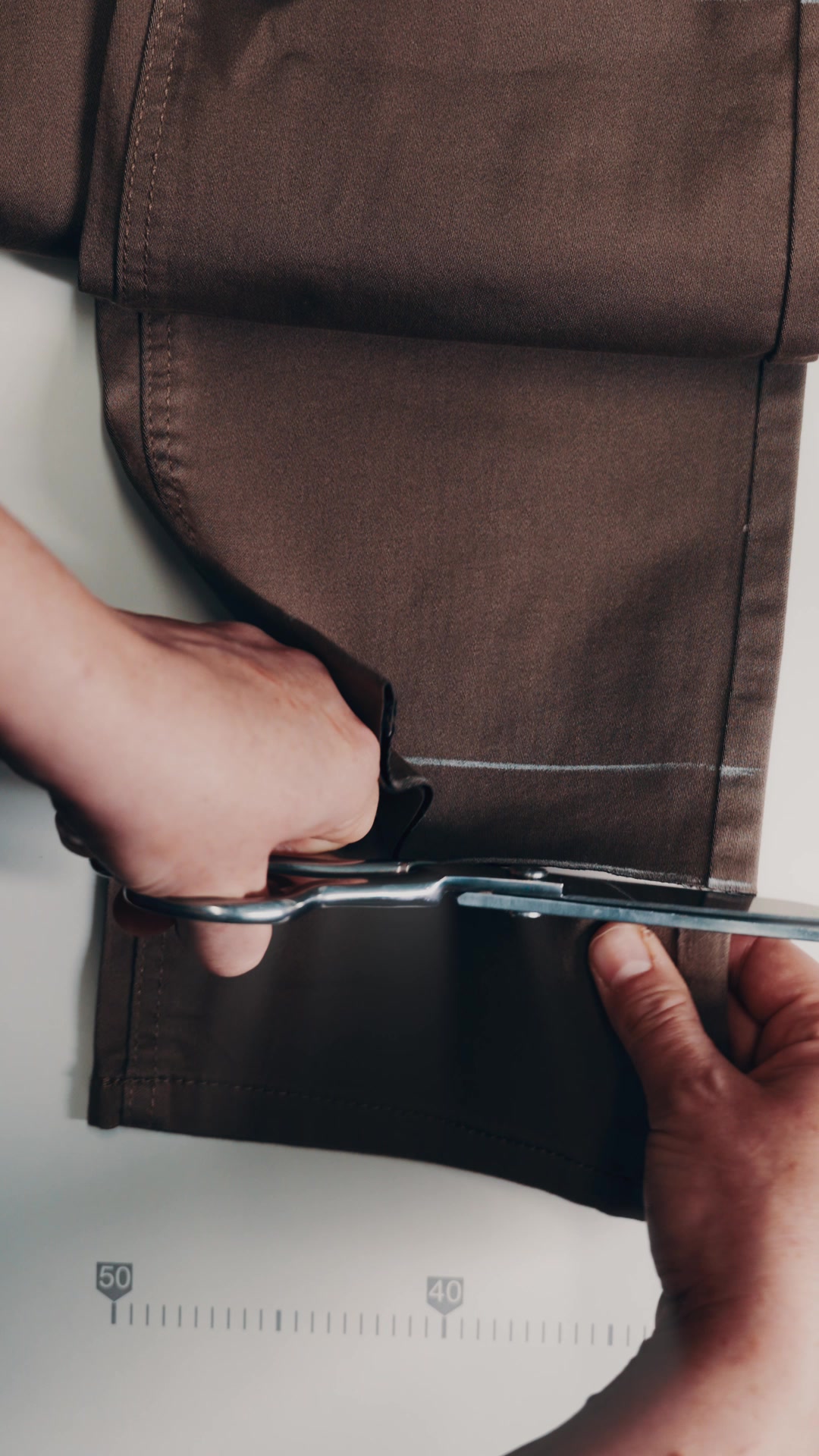 Tailor's hands hemming a pair of brown dress trousers with shears